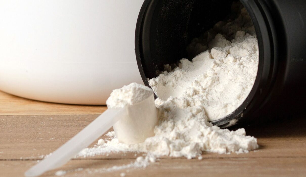 Creatine for muscle gain powder spilling from black container with measuring scoop on wooden surface